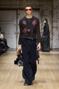 Male model walks a fashion runway wearing a brown knit cardigan with red and black patch squares, red sunglasses, and layered necklaces. He carries a cluster of fluffy accessories.