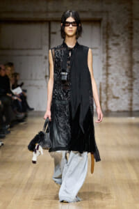 Runway model in a black sequined, mixed-texture outfit with a long scarf and sunglasses, carrying a black handbag with a plush stuffed animal hanging from it.