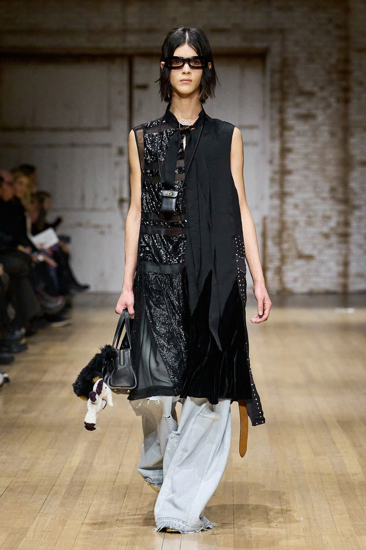Runway model in a black sequined, mixed-texture outfit with a long scarf and sunglasses, carrying a black handbag with a plush stuffed animal hanging from it.