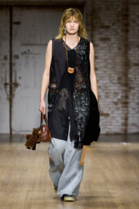Model walking a runway in a black layered outfit with sheer lace panels and oversized light-wash jeans, carrying a brown mini bag and a teddy bear keychain