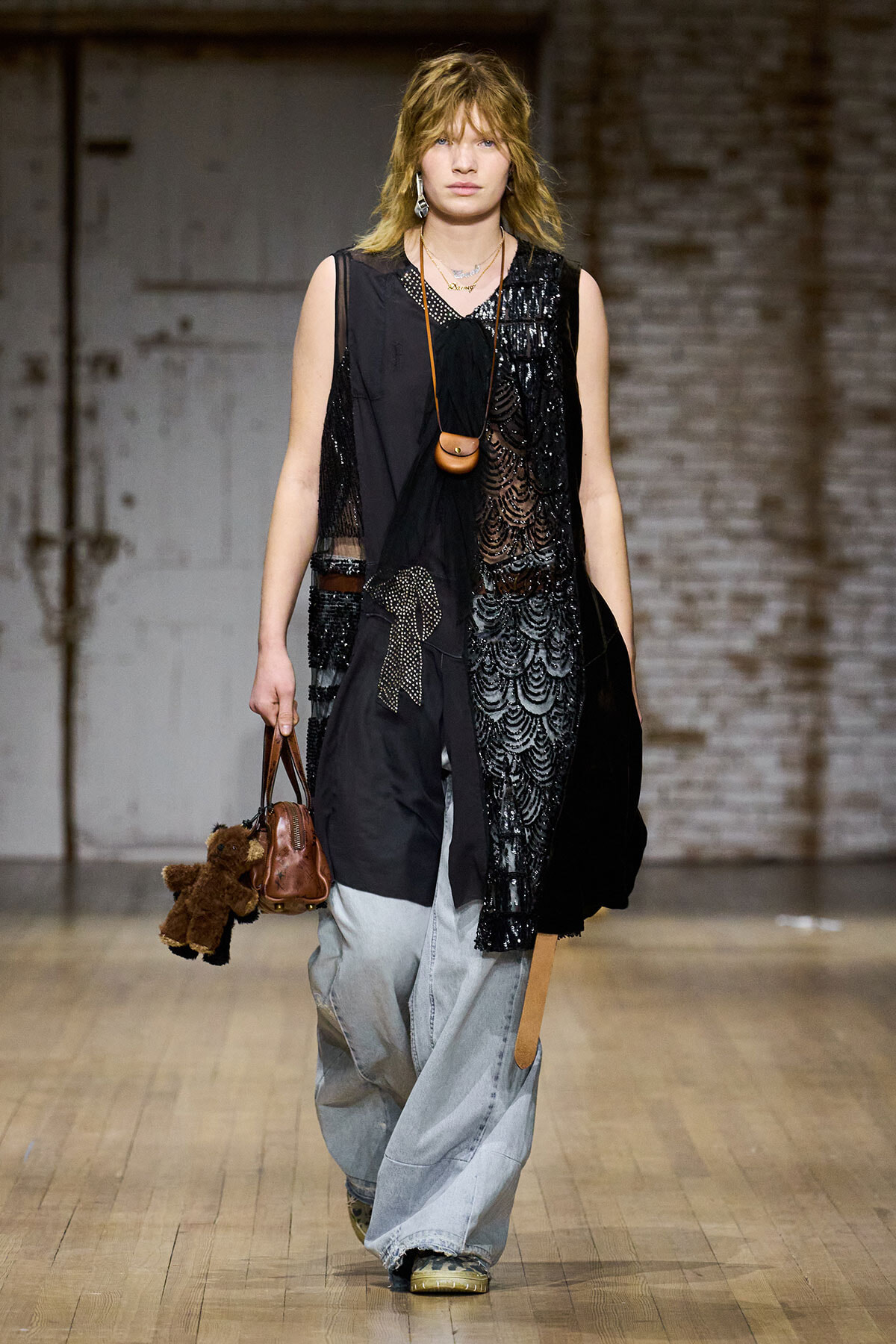 Model walking a runway in a black layered outfit with sheer lace panels and oversized light-wash jeans, carrying a brown mini bag and a teddy bear keychain