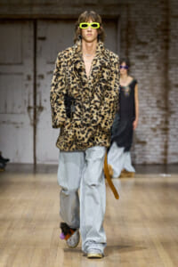 Model walking a fashion runway in a leopard-print faux-fur coat, oversized light-wear jeans, and neon yellow sunglasses; accessorized with bold jewelry and a black handbag.