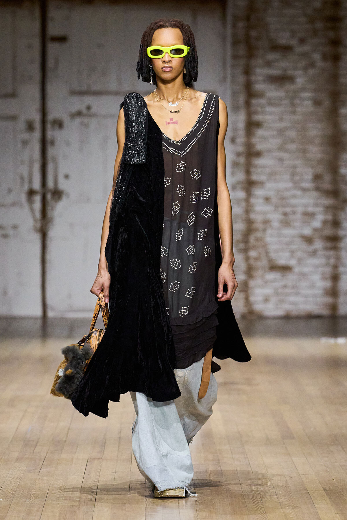 Model on a runway wearing a sheer black beaded dress with geometric patterns, neon lime sunglasses, and a black velvet scarf draped over the shoulder, carrying a fur-accented bag and light-blue jeans underneath.