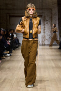 Model walking a fashion runway in a tan leather vest, wide brown pants, and orange sunglasses, with a matching belt and accessories.