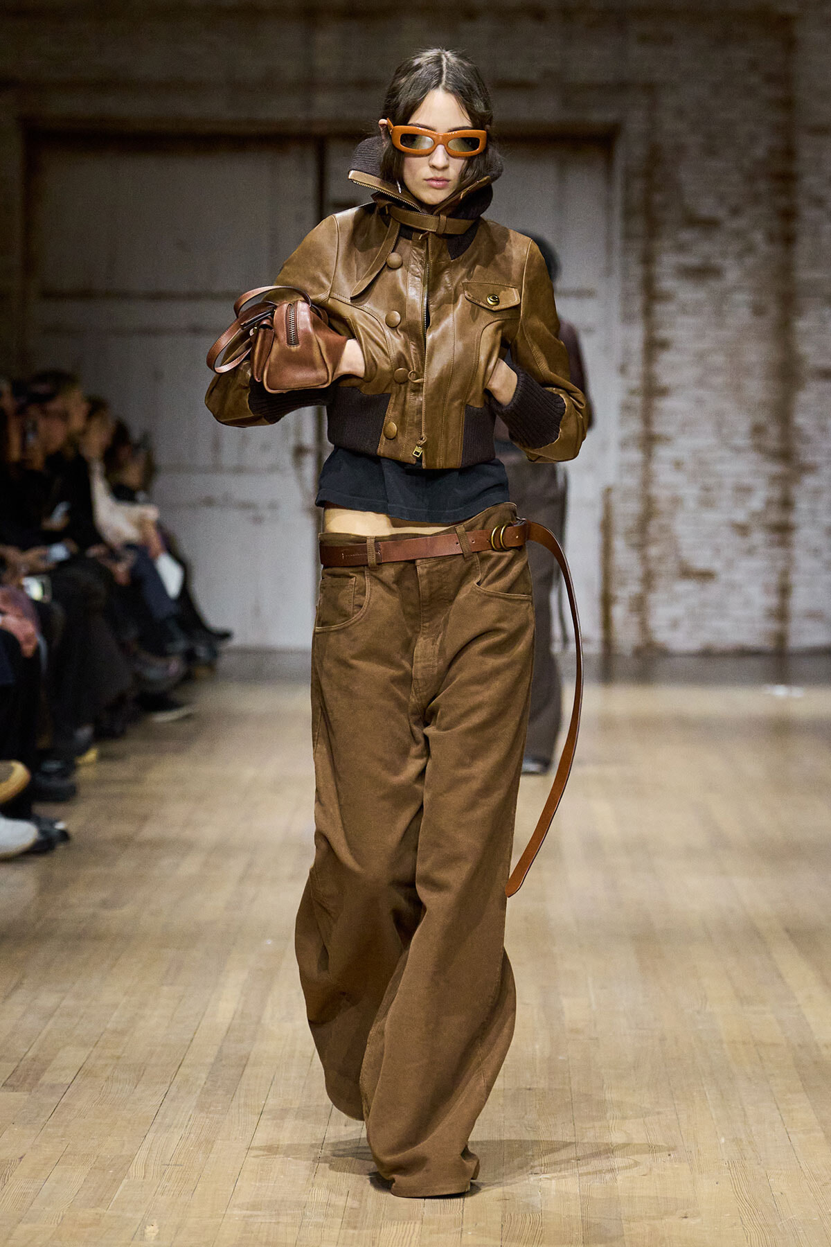 Model walking a runway in a brown leather cropped jacket, matching bag, and wide-leg brown trousers with orange sunglasses.