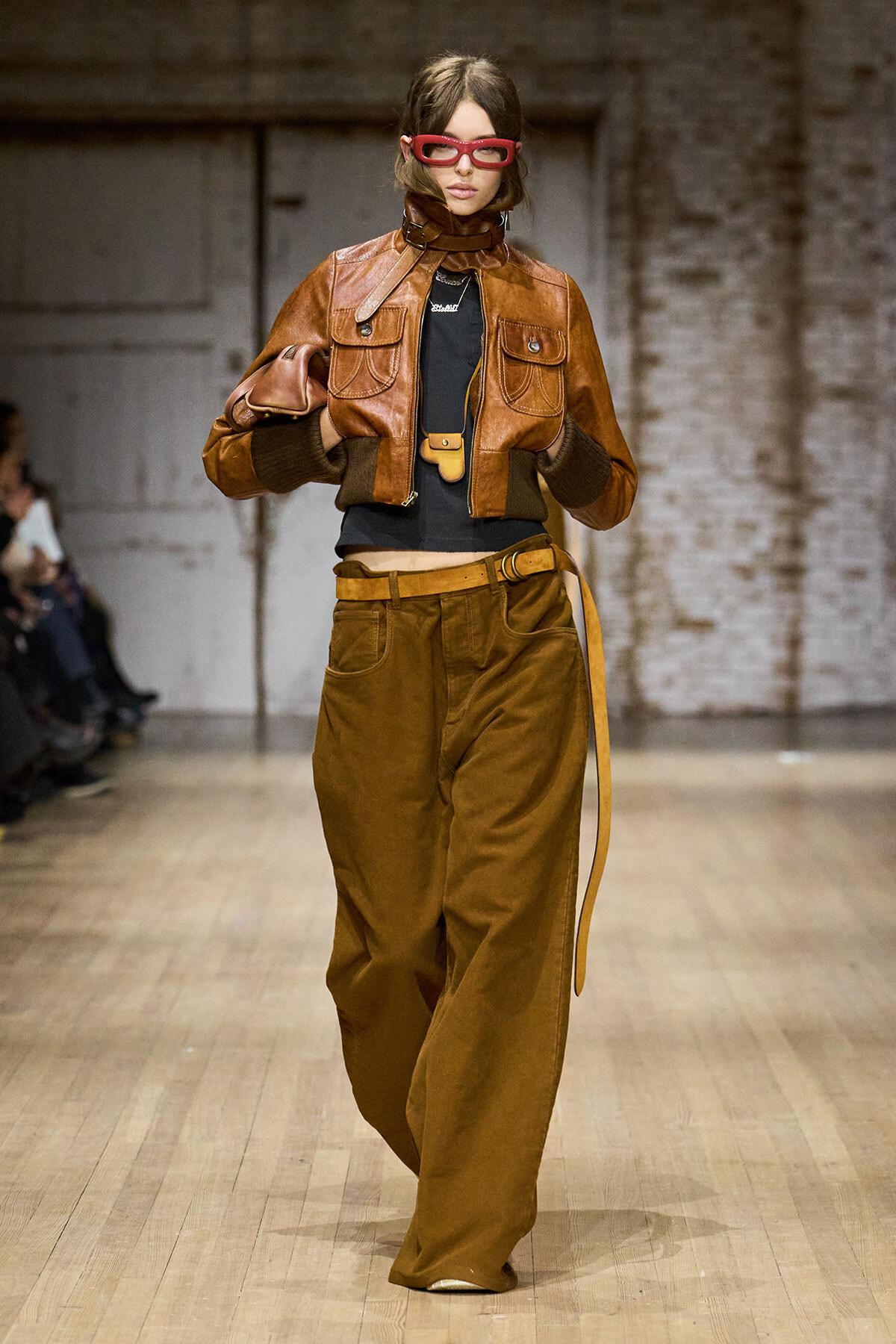 Model walks the runway in a brown leather cropped jacket, wide tan pants, and red rectangular glasses on a fashion show catwalk.
