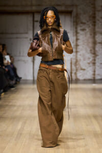 Model on a fashion runway wearing a brown leather vest over a black top, wide brown pants, a brown belt, orange sunglasses, and carrying a brown bag under her arm.