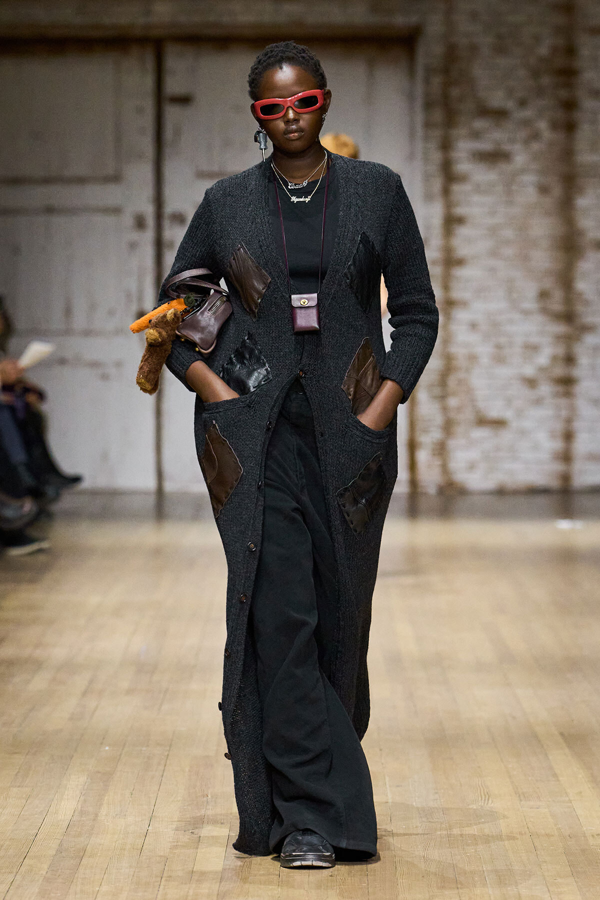 Model walking runway in a long dark cardigan with brown patchwork, black outfit, red sunglasses, and layered necklaces; carrying stuffed items under arm and small crossbody bag.