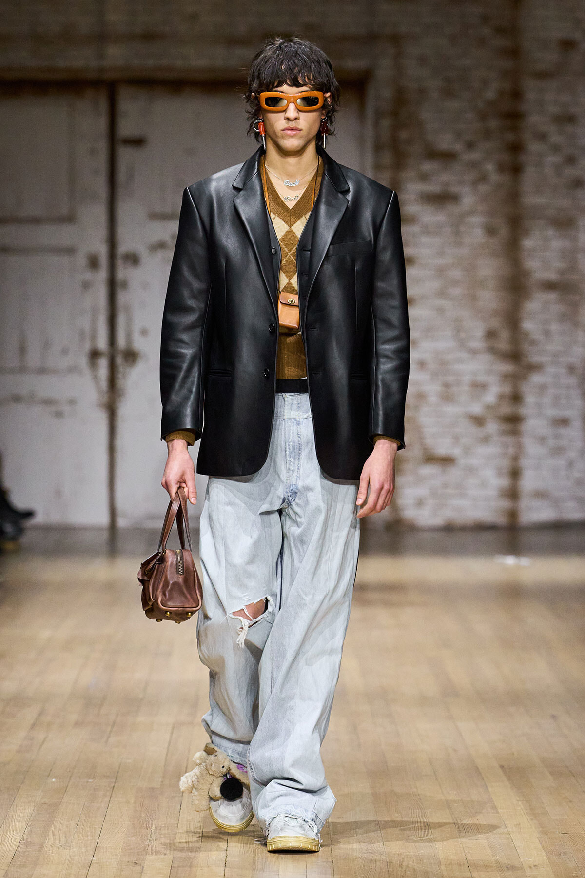 Male model on a runway wearing a black leather blazer over a tan argyle sweater, oversized light-blue ripped jeans, orange sunglasses, and carrying a brown handbag.