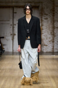 Runway model wearing a black blazer over a vest with oversized light jeans and large plush teddy-bear slippers, sunglasses, and a small bag on a wooden runway floor.