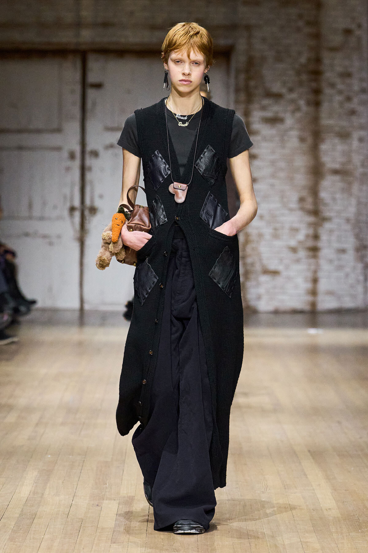 Runway model walks in a long black knitted vest with oversized patch pockets over a dark outfit, short red hair, accessorized with a pendant necklace and earrings, and carrying a brown bag with a plush toy accessory.