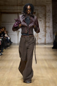 Model walking runway in a brown leather bomber jacket, dark top, and wide-leg brown pants, holding a purple handbag.