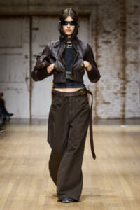 Model walking a fashion runway in a cropped brown leather jacket with oversized sleeves, black top, wide brown trousers, and sunglasses.