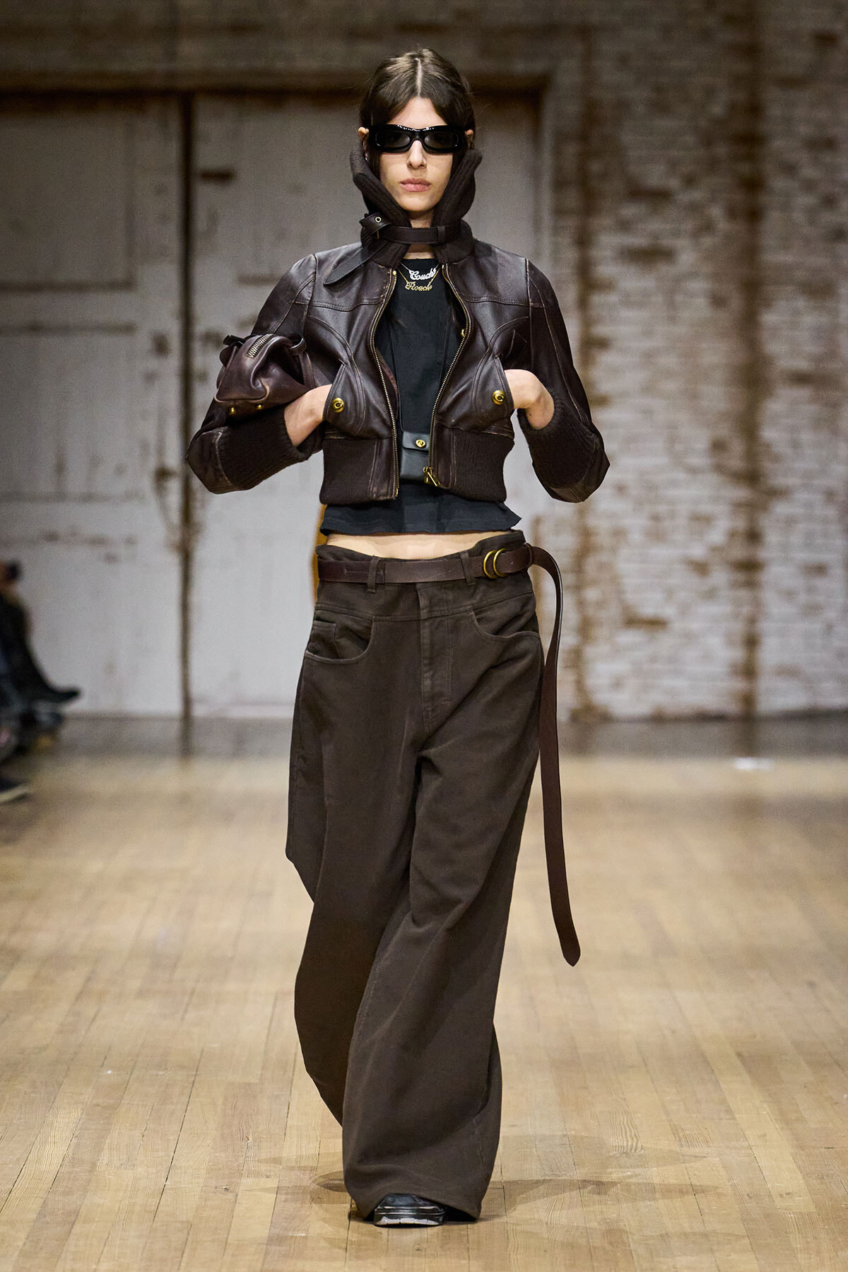 Model walking a fashion runway in a cropped brown leather jacket with oversized sleeves, black top, wide brown trousers, and sunglasses.