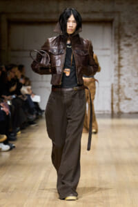 Model walks a runway in a dark brown leather bomber jacket, black top, and oversized brown trousers with a belt and small pouch.