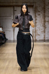 Model walking a fashion runway in a dark, cropped black top layered with a brown leather padded vest and oversized black wide-leg pants, accessorized with a belt and a small brown bag under her arm.