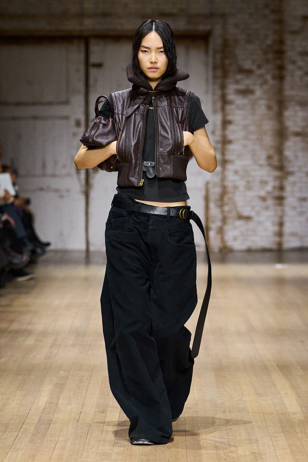 Model walking a fashion runway in a dark, cropped black top layered with a brown leather padded vest and oversized black wide-leg pants, accessorized with a belt and a small brown bag under her arm.