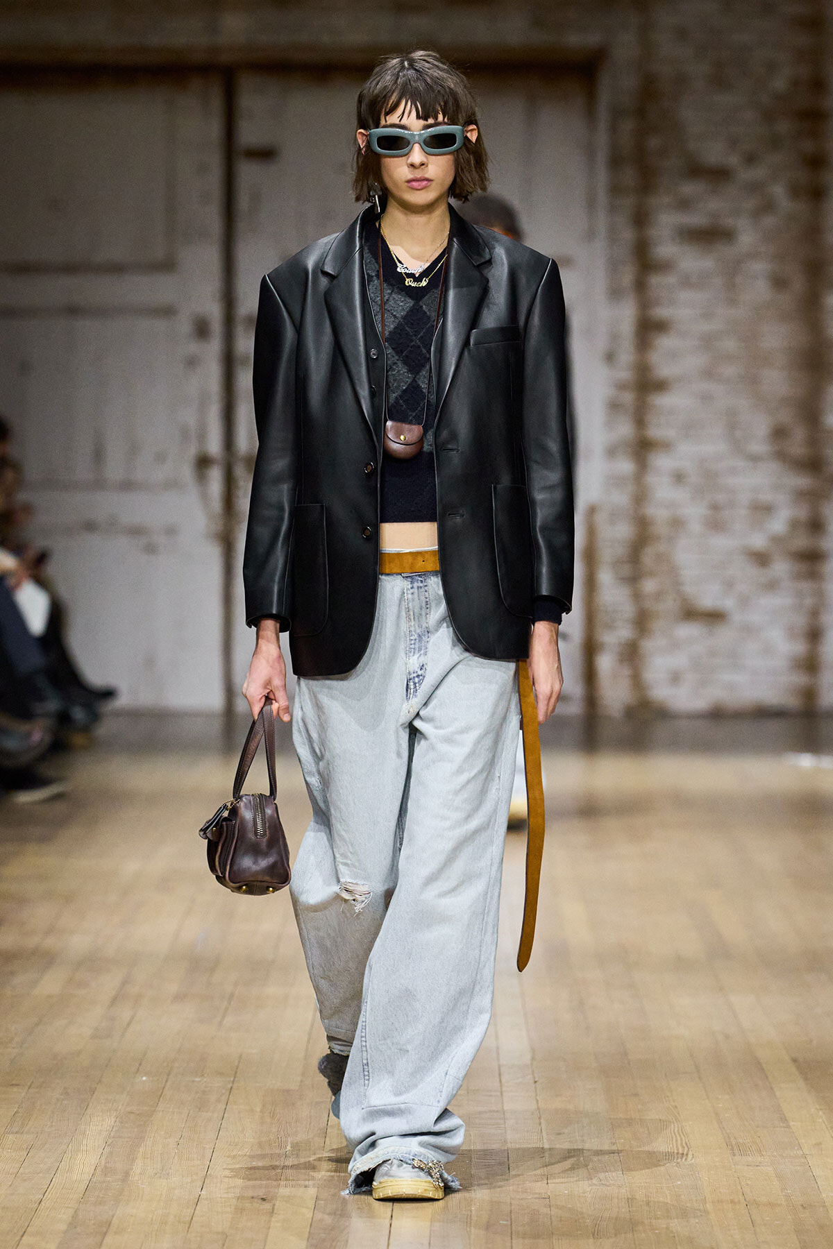 Model wearing oversized light-wash jeans, black leather blazer, cropped sweater, sunglasses, and a small brown handbag on a runway.