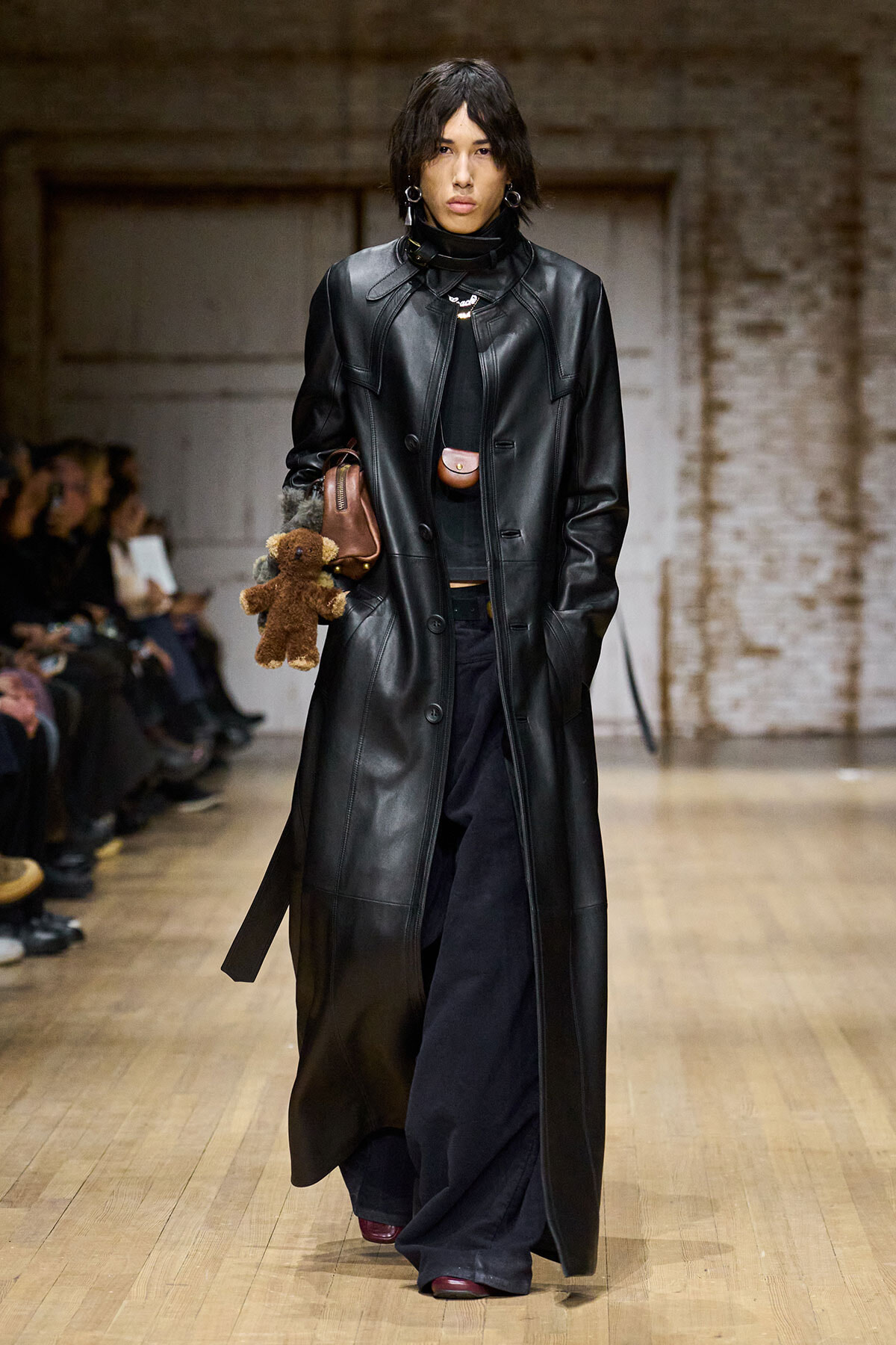 Model walking a runway in a long black leather trench, holding a brown purse and teddy bear accessory, with dark wide-leg pants and red shoes visible.