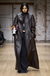 Model walks the runway in a long dark brown leather coat, black wide-leg pants, and a plush dog-themed handbag/purse tucked under her arm.
