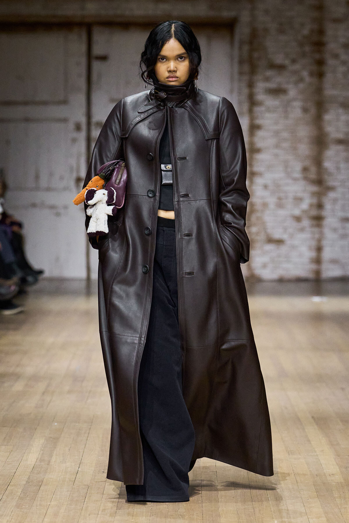 Model walks the runway in a long dark brown leather coat, black wide-leg pants, and a plush dog-themed handbag/purse tucked under her arm.