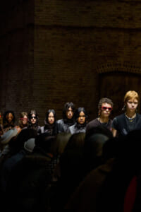 Models in dark outfits lined up on a dim runway against a brick wall, with audience silhouettes in the foreground.