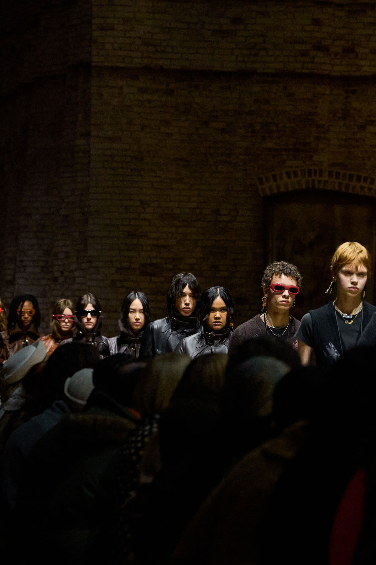 Models in dark outfits lined up on a dim runway against a brick wall, with audience silhouettes in the foreground.