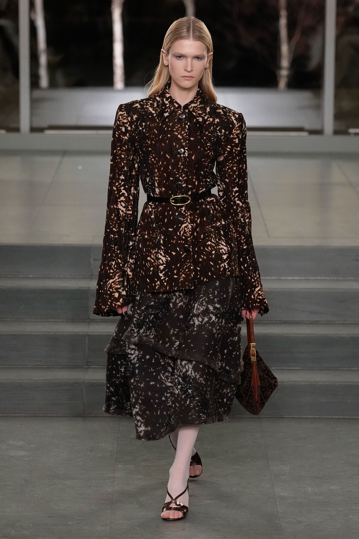 Model on a runway wearing a brown and cream speckled blouse with a matching tiered skirt, a black belt, and a brown patterned handbag.
