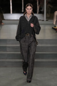Model walking runway in a dark textured grey pantsuit with a loose cardigan, wearing a black belt and carrying a black shoulder bag.