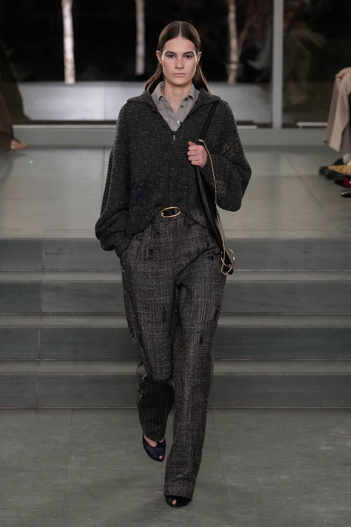 Model walking runway in a dark textured grey pantsuit with a loose cardigan, wearing a black belt and carrying a black shoulder bag.