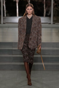 Model on a runway wearing a brown plaid blazer over a dark knit top and a textured tweed skirt, brown knee-high boots, and a tan clutch.