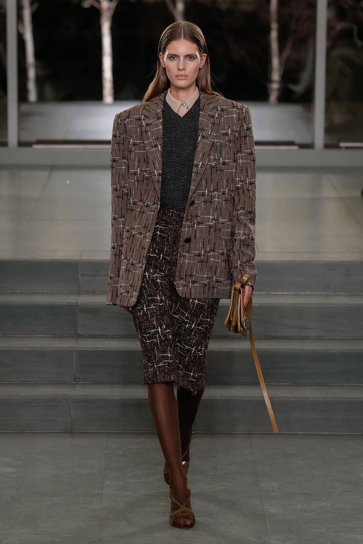Model on a runway wearing a brown plaid blazer over a dark knit top and a textured tweed skirt, brown knee-high boots, and a tan clutch.