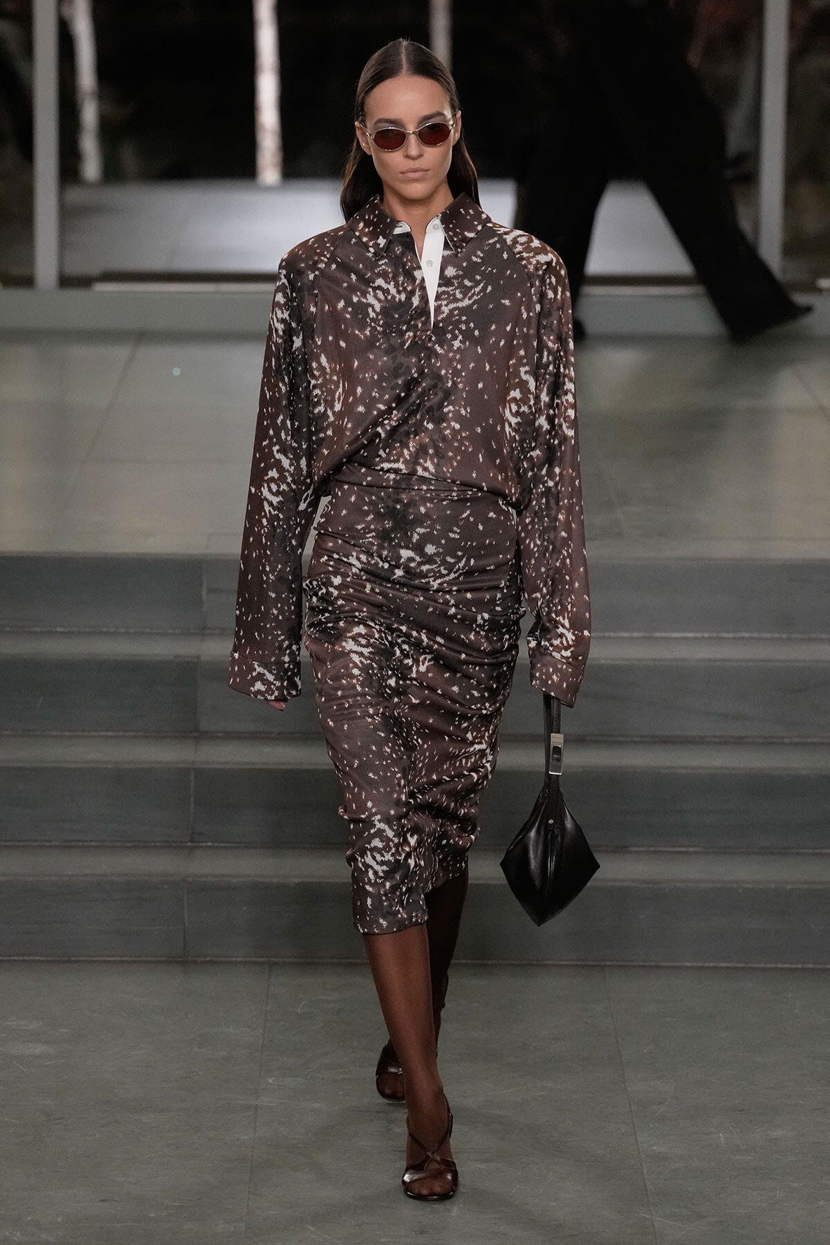 Model walks a runway in a brown speckled midi dress with a white collar, sunglasses, and a small black handbag.