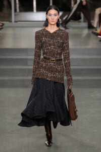 Runway model wearing a textured brown tweed top with a belt and a voluminous navy skirt, carrying a brown handbag.