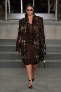 Model walks a runway in a dark brown, splattered-pattern coat over a brown outfit, wearing oval sunglasses and carrying a black bag.