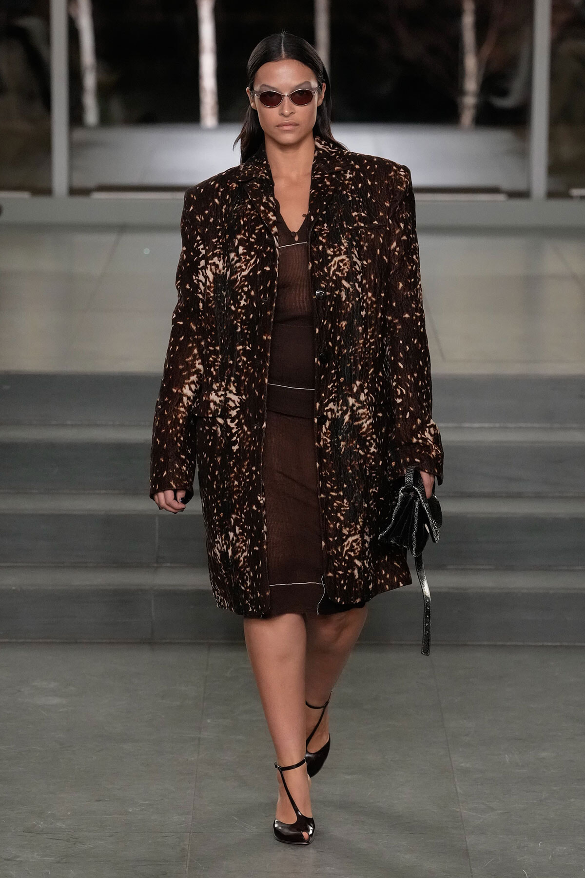 Model walks a runway in a dark brown, splattered-pattern coat over a brown outfit, wearing oval sunglasses and carrying a black bag.