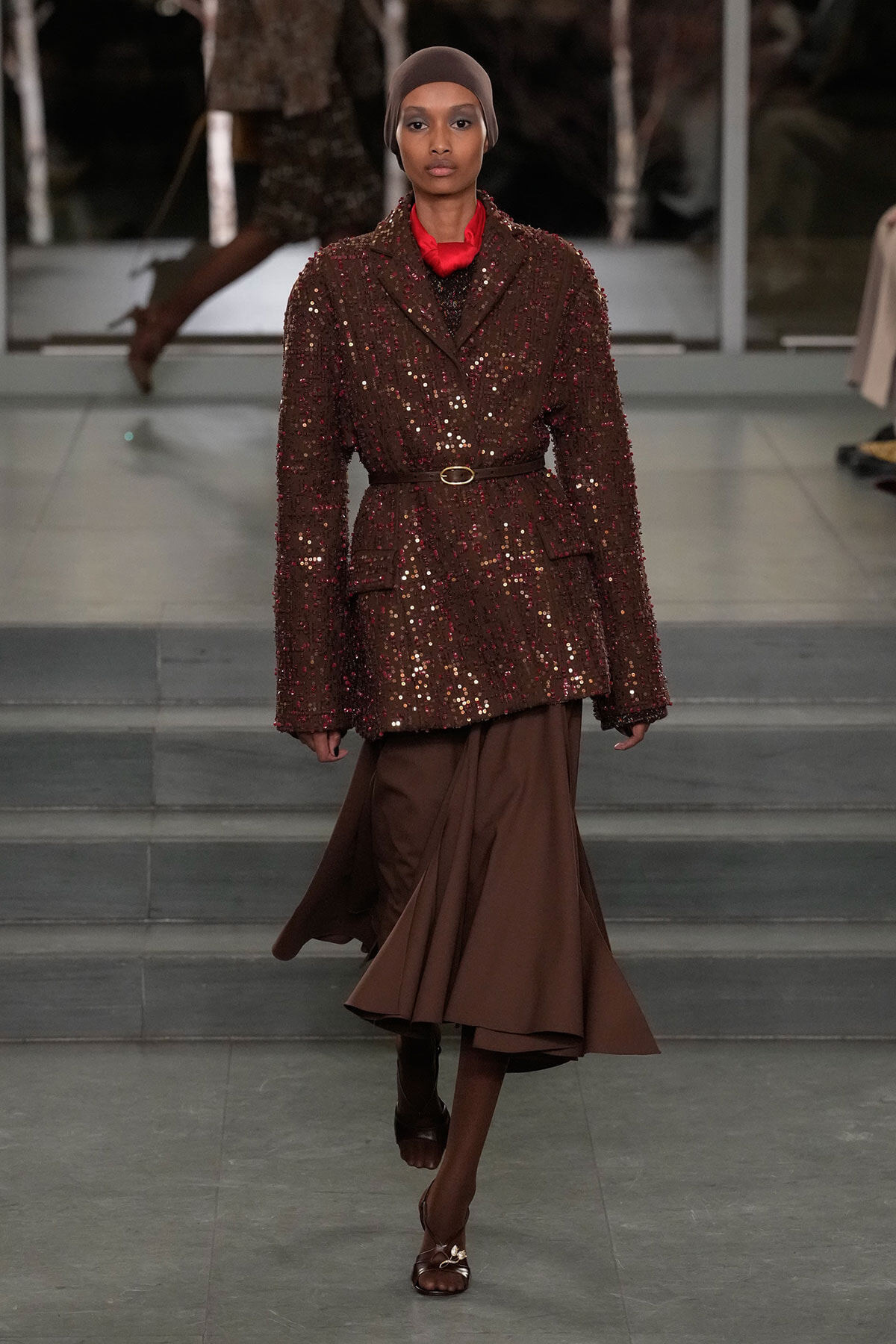 Model walking a fashion runway in a brown sequin blazer belted at the waist, paired with a flowing brown midi skirt and taupe headscarf.
