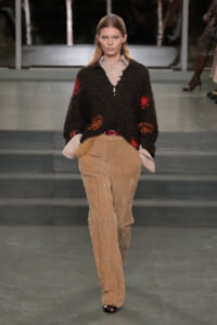 Model walks the runway wearing a loose black knit sweater with red and orange patches over a beige button-up shirt and tan corduroy pants with a red belt.