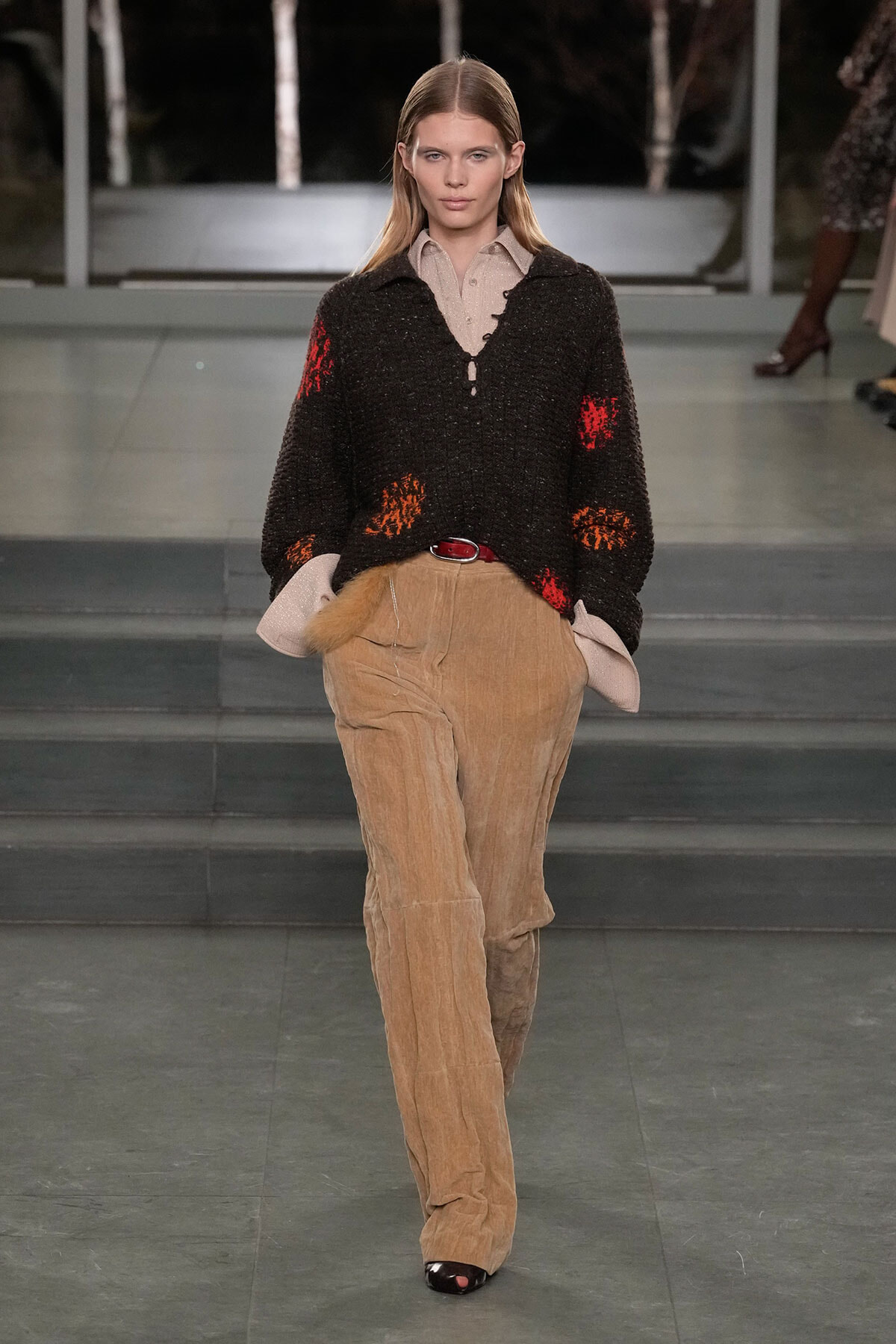 Model walks the runway wearing a loose black knit sweater with red and orange patches over a beige button-up shirt and tan corduroy pants with a red belt.