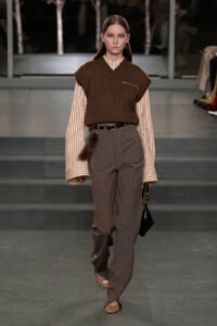 Female model walks a runway in a brown knit vest over a striped button-down shirt paired with brown plaid trousers and a slim belt, carrying a small black bag.