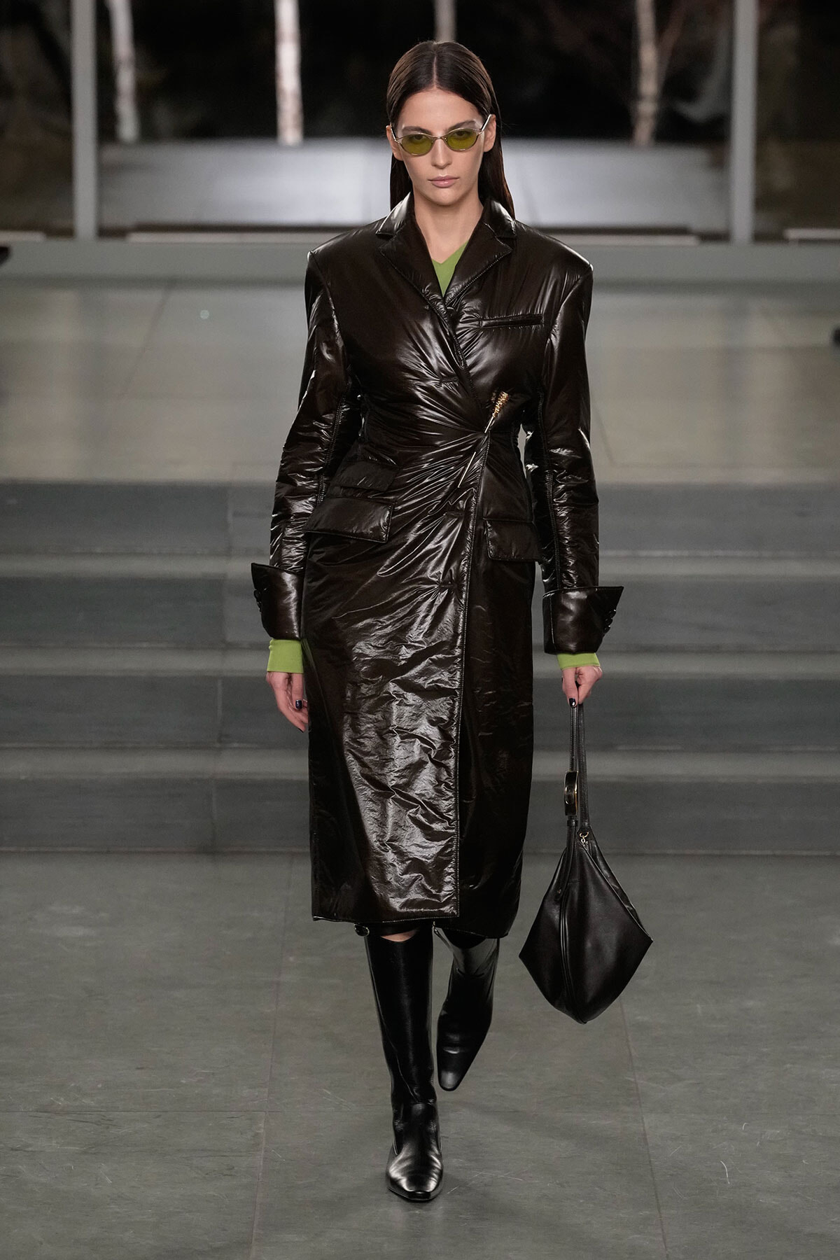 Model on a runway wearing a shiny black leather trench, green top peeking out, and yellow-tinted sunglasses with a black handbag.