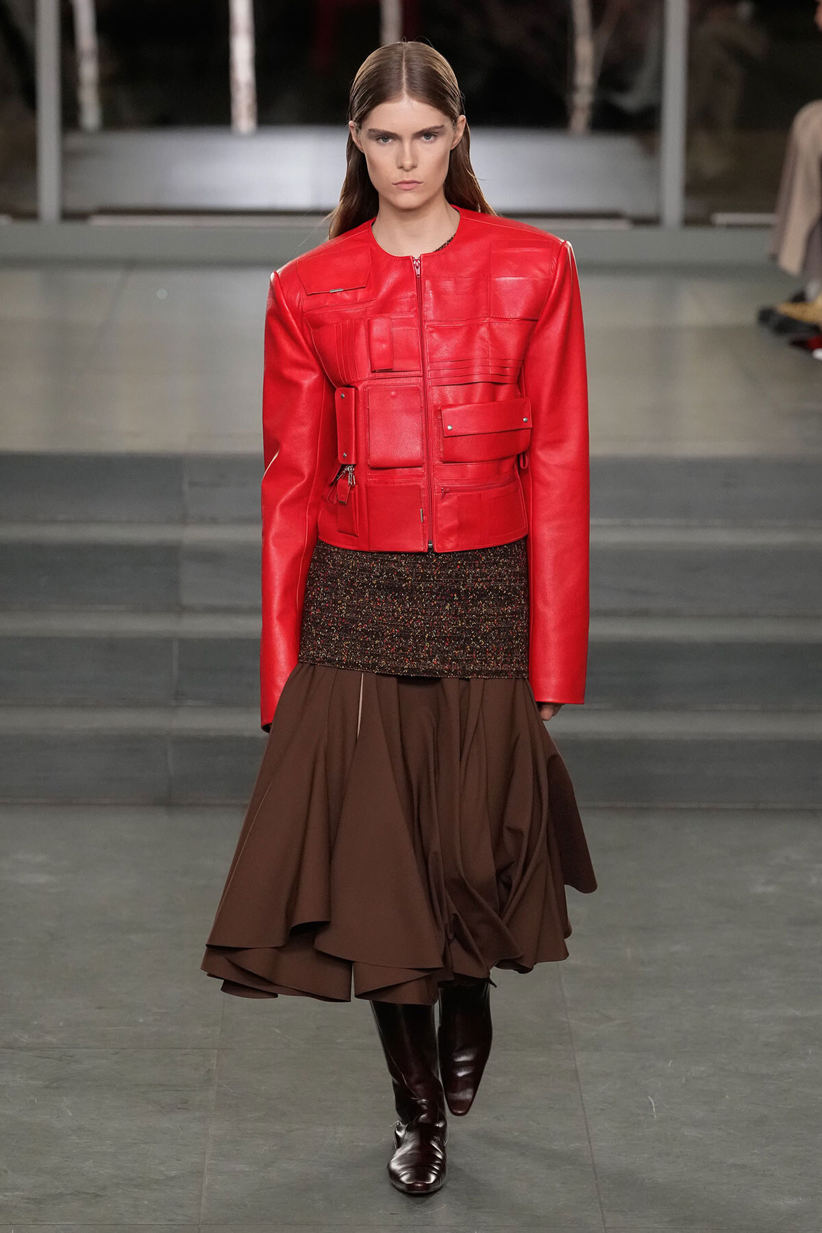 Model walking a runway in a bright red leather utility jacket with multiple pockets, paired with a textured black top and a voluminous brown layered skirt, knee-high boots.