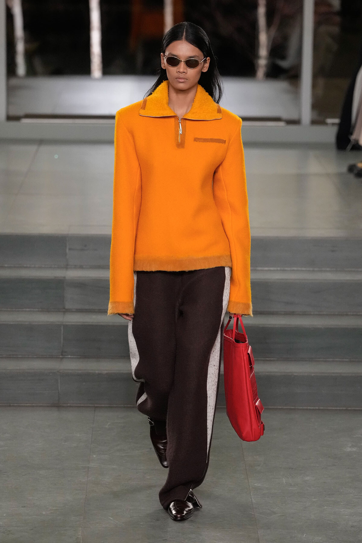 Model walking runway in a bright orange fleece top with a zip collar, brown wide-leg pants with white side panels, and black loafers, carrying a red handbag with sunglasses on.