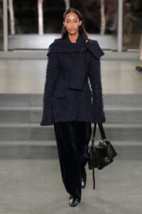 Model on a runway wearing a navy textured coat with a draped navy scarf and a black leather handbag, accessorized in dark tones.