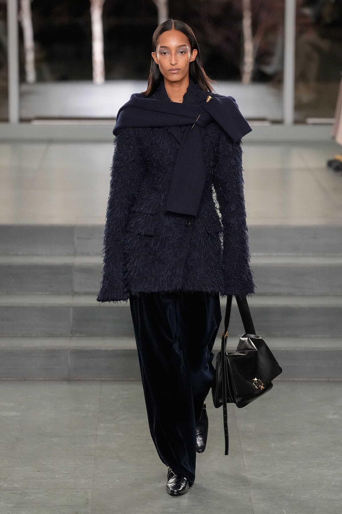 Model on a runway wearing a navy textured coat with a draped navy scarf and a black leather handbag, accessorized in dark tones.