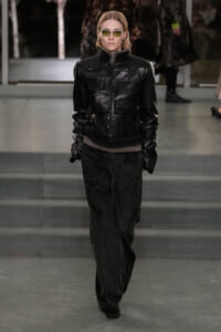 Model walking a runway in a black leather jacket with a fur collar, dark pants, and yellow-green sunglasses.