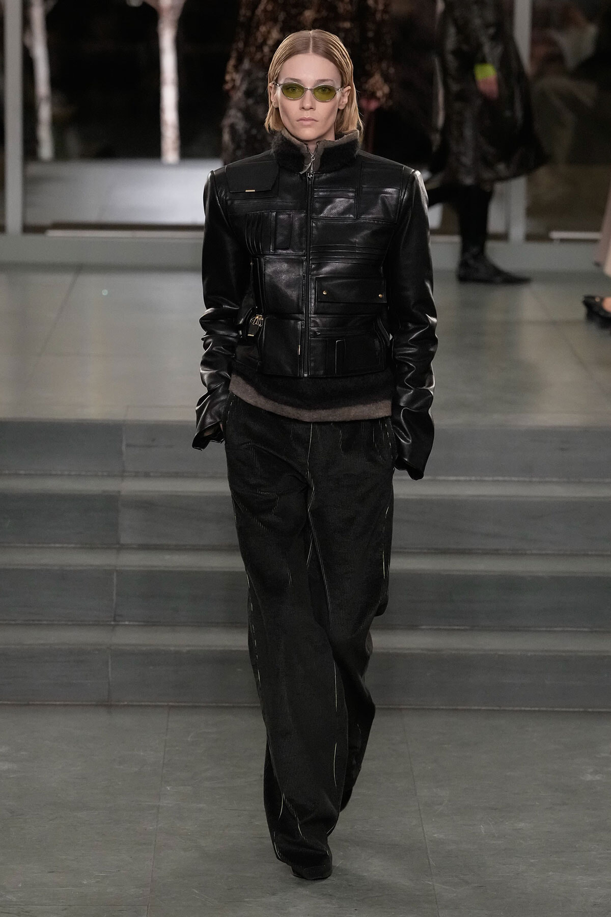 Model walking a runway in a black leather jacket with a fur collar, dark pants, and yellow-green sunglasses.