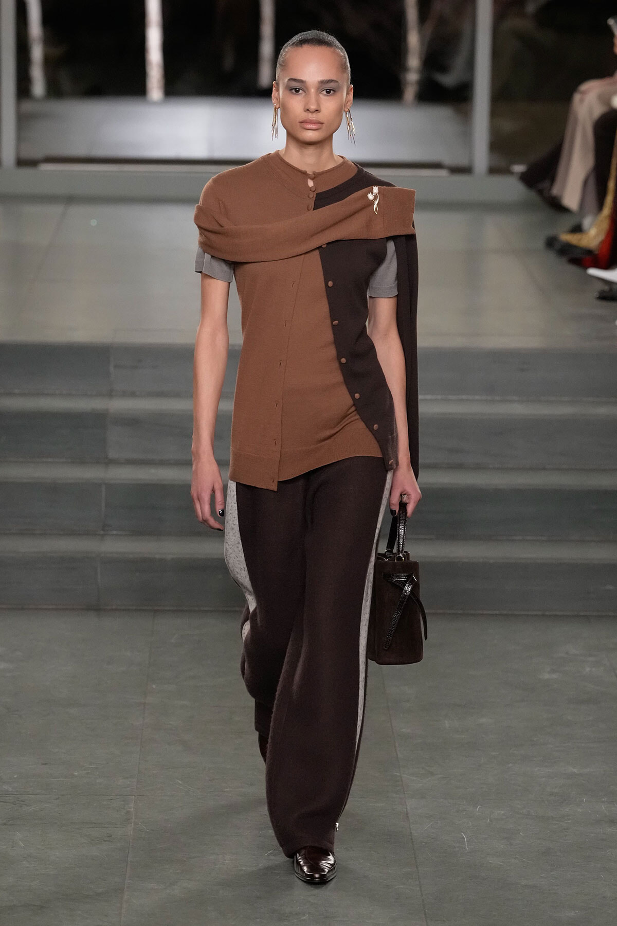 Model walking a runway in a brown asymmetrical tunic layered over a gray top, with dark trousers that have gray side panels and a small black cape draped over one shoulder.