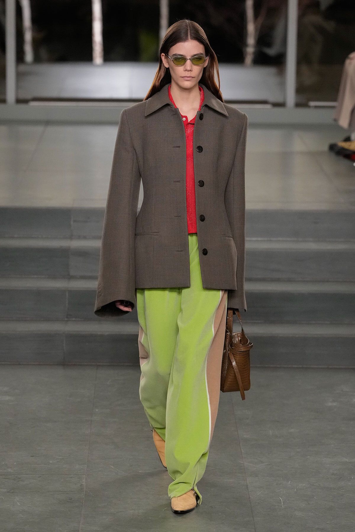 Model walking runway in oversized gray coat over red top and lime green pants with beige panels, carrying a brown bag and wearing yellow-tinted sunglasses.