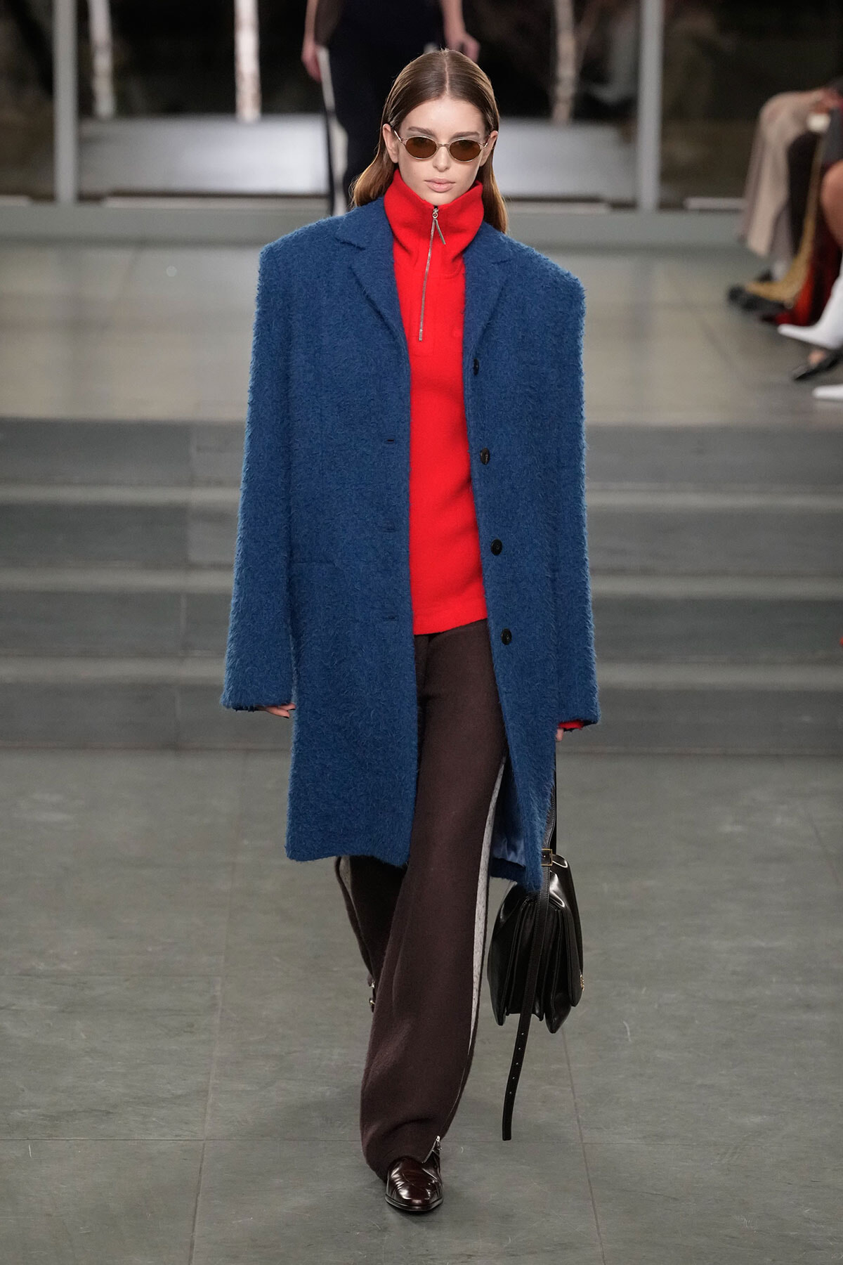 Model walks a runway in a bold blue textured overcoat over a bright red zip-neck sweater, wearing brown wide-leg pants and sunglasses, carrying a black handbag.